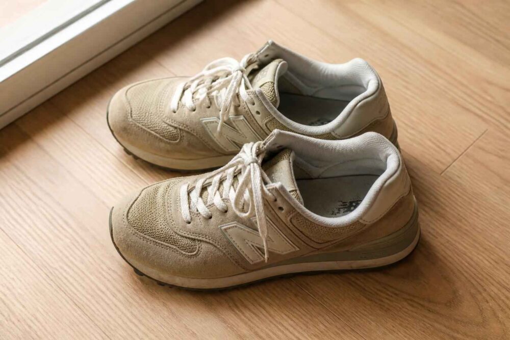 Dwie jasnobeżowe pary sneakersów New Balance Dwa jasnobeżowe sneakersy New Balance leżące na drewnianej podłodze przy oknie