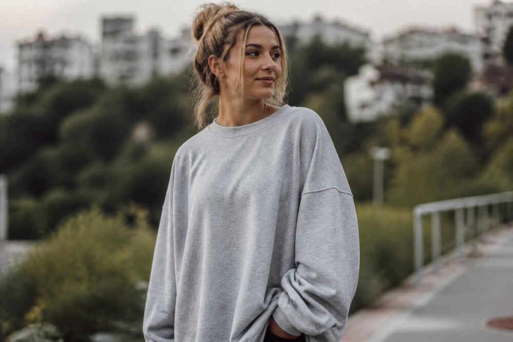Młoda kobieta w jasnej bluzie na tle miejskiego parku Kobieta w jasnoszarej bluzie stoi przy ścieżce, a za nią widać drzewa i miejskie budynki